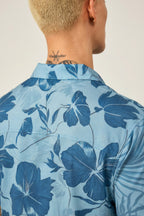 A person with short, light blond hair and a neck tattoo is seen from behind wearing the 686 Men's Nomad Perforated Button Down Shirt by 686, featuring a lightweight, moisture-wicking fabric and a dark blue floral pattern.