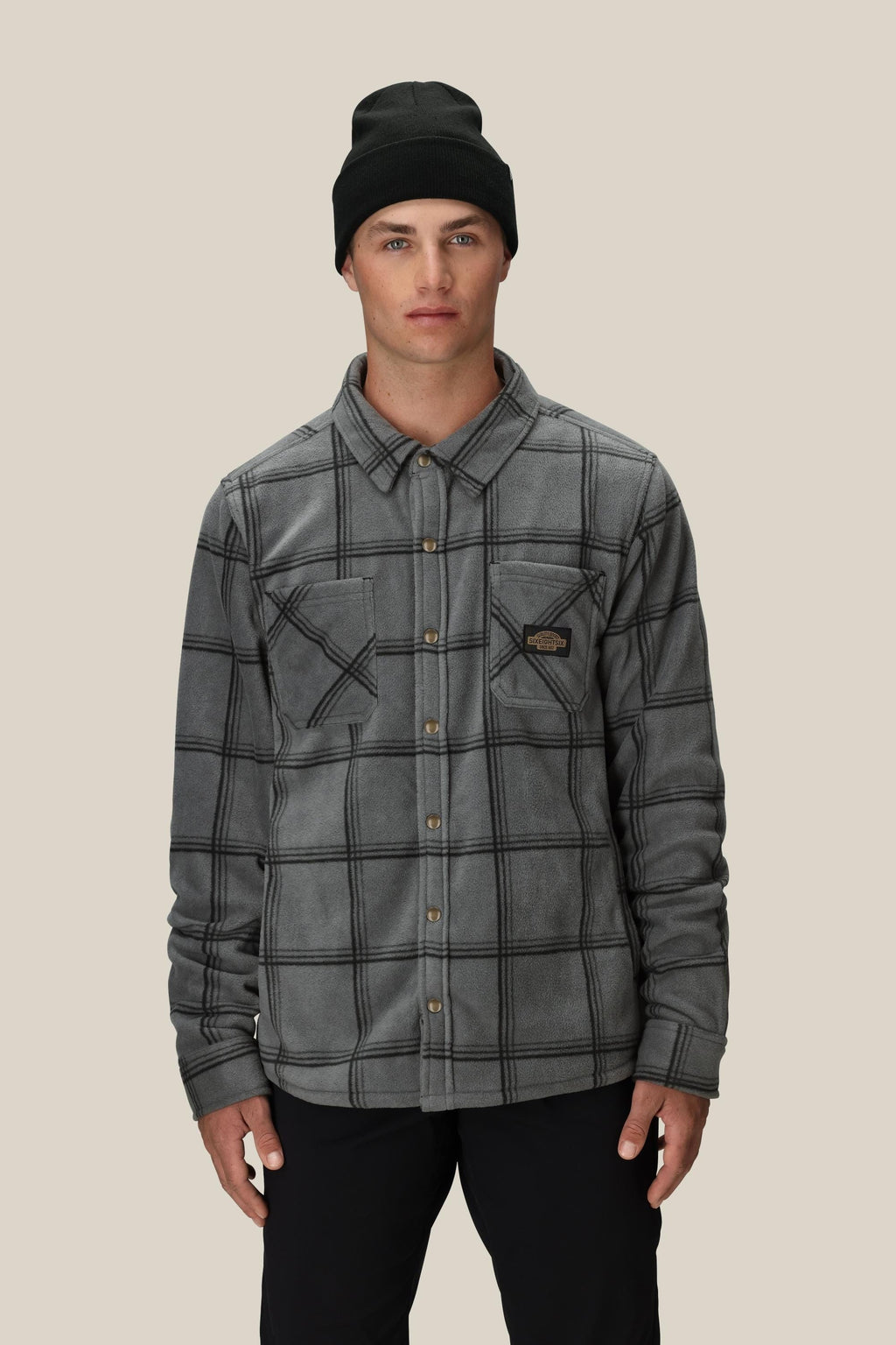 A young man in the 686 Mens Sierra Fleece Flannel by 686 and a black beanie stands against a light beige background, looking at the camera. This gray plaid shacket is an ideal layering piece for spring riding.