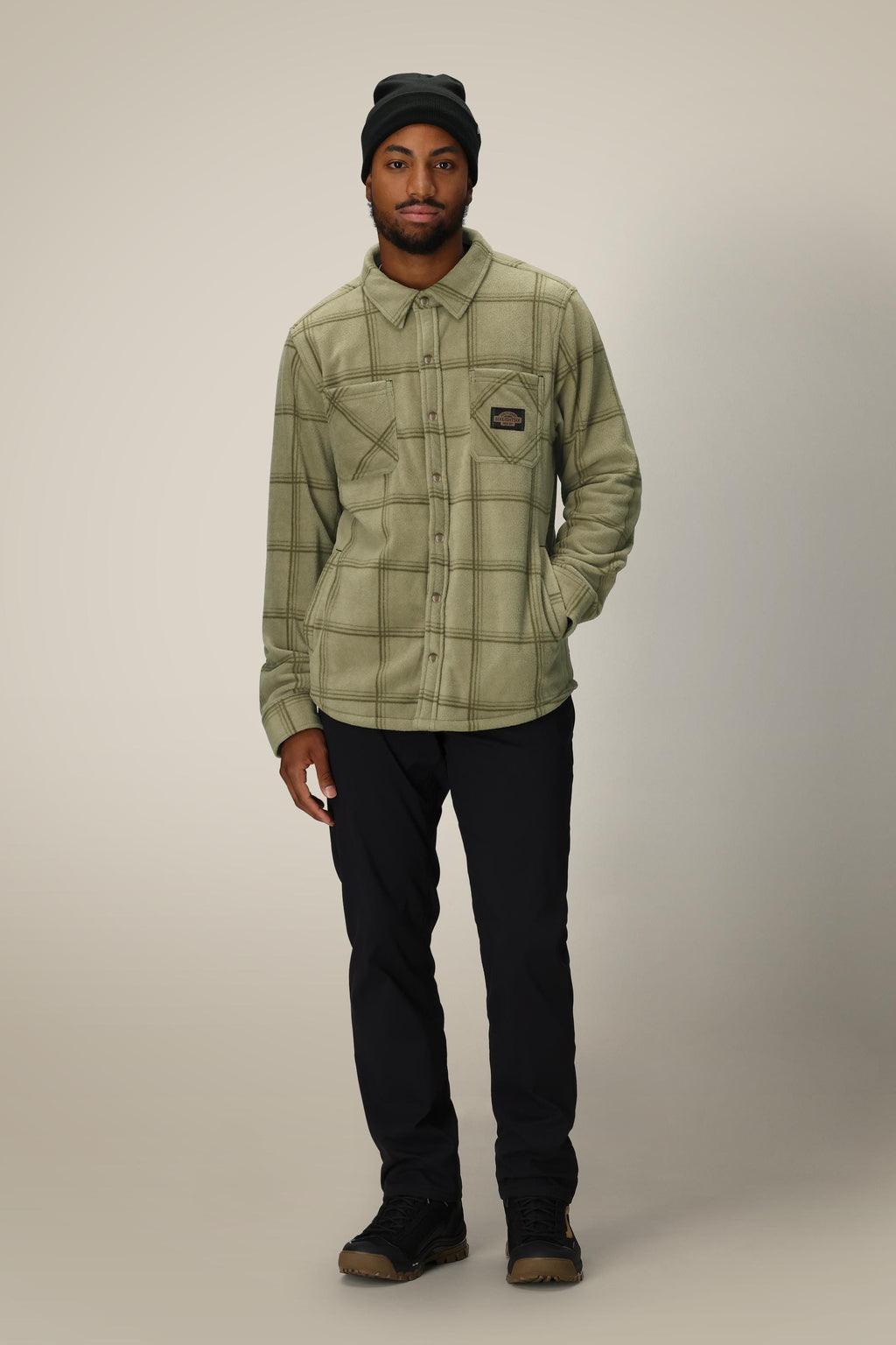 A man wearing a 686 Mens Sierra Fleece Flannel in light green plaid, a black beanie, black pants, and brown shoes stands against a plain light background with one hand in his pocket.