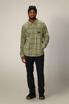 A man wearing a 686 Mens Sierra Fleece Flannel in light green plaid, a black beanie, black pants, and brown shoes stands against a plain light background with one hand in his pocket.