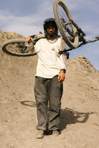 A person in 686 Cruiser Pant - Wide Fit by 686 stands on a rocky slope beneath a partly cloudy sky, wearing a helmet and carrying a mountain bike over their shoulder.