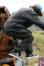 A person in a black jacket, 686 Cruiser Pant - Wide Fit by 686, Vans sneakers, and a blue cap crouches on a rusted graffiti-covered structure outdoors with green hills in the background. Their face is turned away.