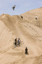 A group of cyclists on a dirt trail, wearing 686 Cruiser Pant - Wide Fit by 686, watch as another cyclist performs a high jump off a rocky hill, with barren terrain and a few onlookers in the background.