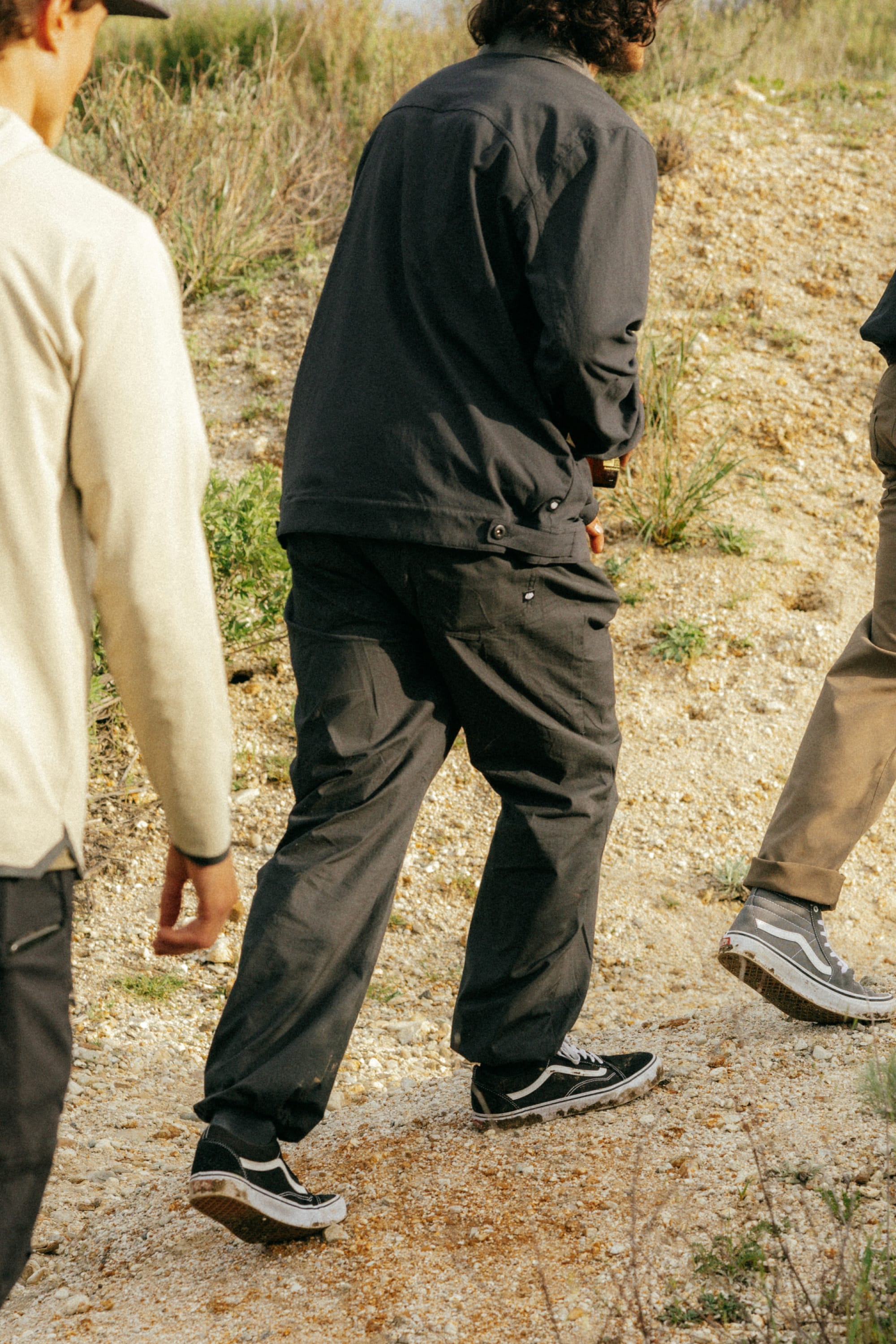 Three people wearing 686 Cruiser Pant - Wide Fit by 686 walk outdoors on a rocky path, their lower bodies in focus. Styles feature wide legs and bungee hems, perfect for casual outings surrounded by grass and shrubs.