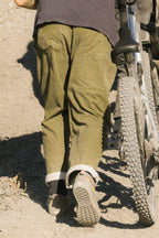 A person wearing 686 Cruiser Pant - Wide Fit by 686 and sneakers walks a mountain bike up a dirt path, seen from behind.
