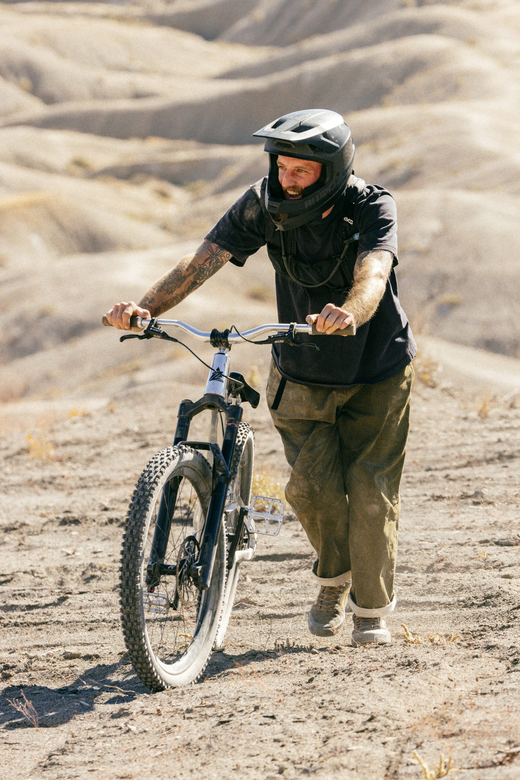 Wearing the 686 Cruiser Pant - Wide Fit by 686, a person in a helmet and protective gear pushes a mountain bike uphill on a dry, rugged trail with sandy hills in the background.