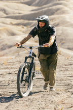 Wearing the 686 Cruiser Pant - Wide Fit by 686, a person in a helmet and protective gear pushes a mountain bike uphill on a dry, rugged trail with sandy hills in the background.