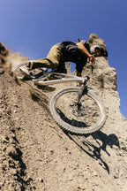 A mountain biker in a helmet and protective gear rides downhill on a steep, rocky slope, kicking up dust in the 686 Cruiser Pant - Wide Fit by 686, with rugged rock formations under a clear blue sky.