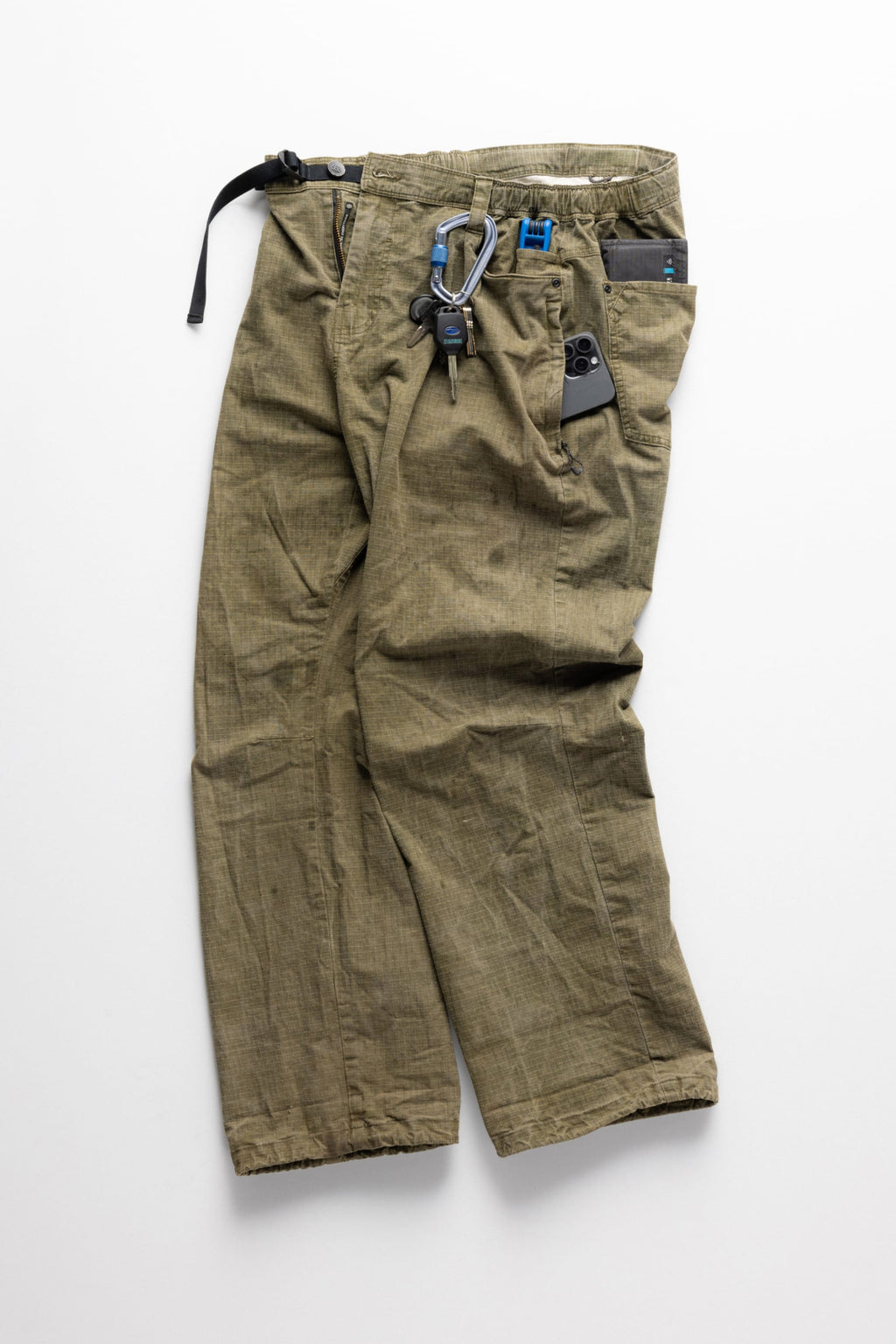 The 686 Cruiser Pant - Wide Fit by 686 in olive green is shown laid flat with a black belt, keychain, and blue carabiner at the waist, plus a black smartphone peeking out of the front pocket, all on a white background.