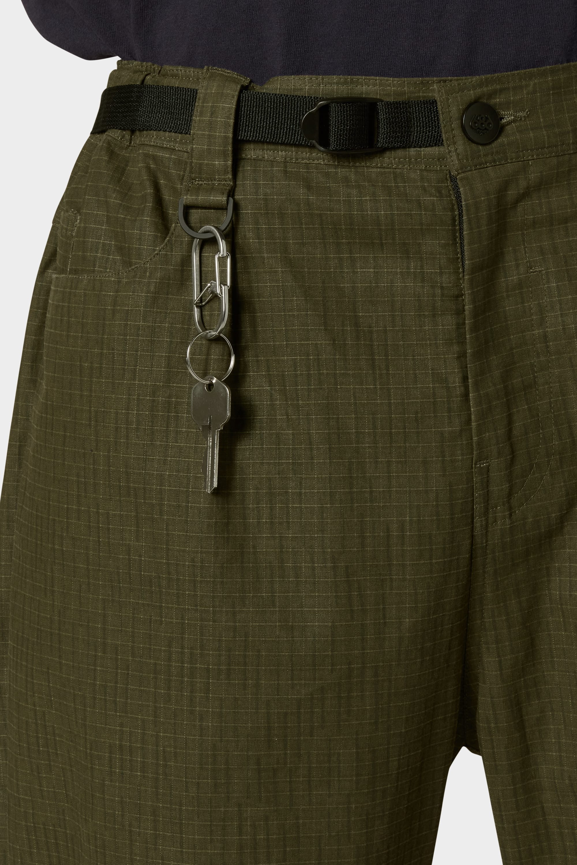 A close-up shows a person wearing the 686 Cruiser Pant - Wide Fit by 686 in olive green ripstop, styled with a black belt and silver keychain on the belt loop, paired with a dark shirt for a modern look.