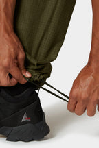 A person cinches the bungee hem at the ankle of 686 Cruiser Pant - Wide Fit by 686 in olive green ripstop, paired with black athletic shoes.