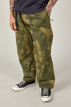 A person wearing loose-fitting green 686 Cruiser Pant - Wide Fit by 686 made from ripstop fabric and black Converse sneakers stands with one hand in a pocket. Their tattooed arms show, paired with a dark shirt.