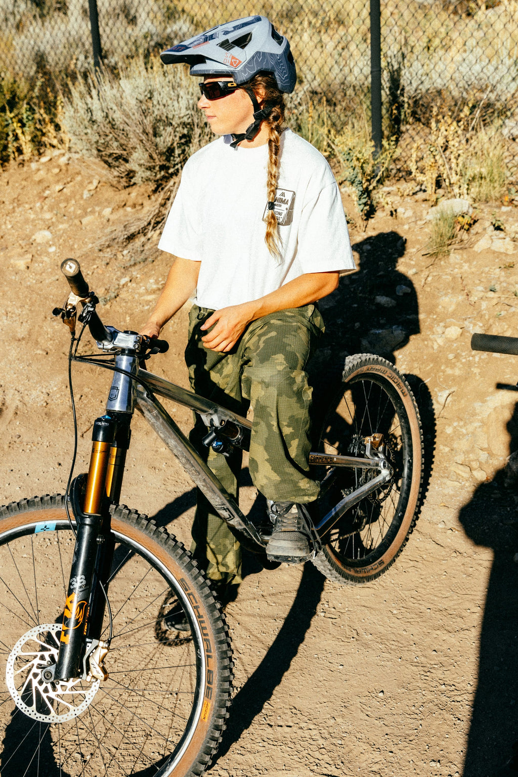 Wearing a helmet, sunglasses, white t-shirt, and the 686 Cruiser Pant - Wide Fit by 686, a person with a long braid sits on a mountain bike beside a wire fence on a dirt path in sunny outdoors.