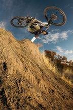 A mountain biker in a helmet and 686 Cruiser Pant - Wide Fit performs an aerial trick over a dirt ramp, tilting the bike sideways with blue sky and autumn foliage behind.