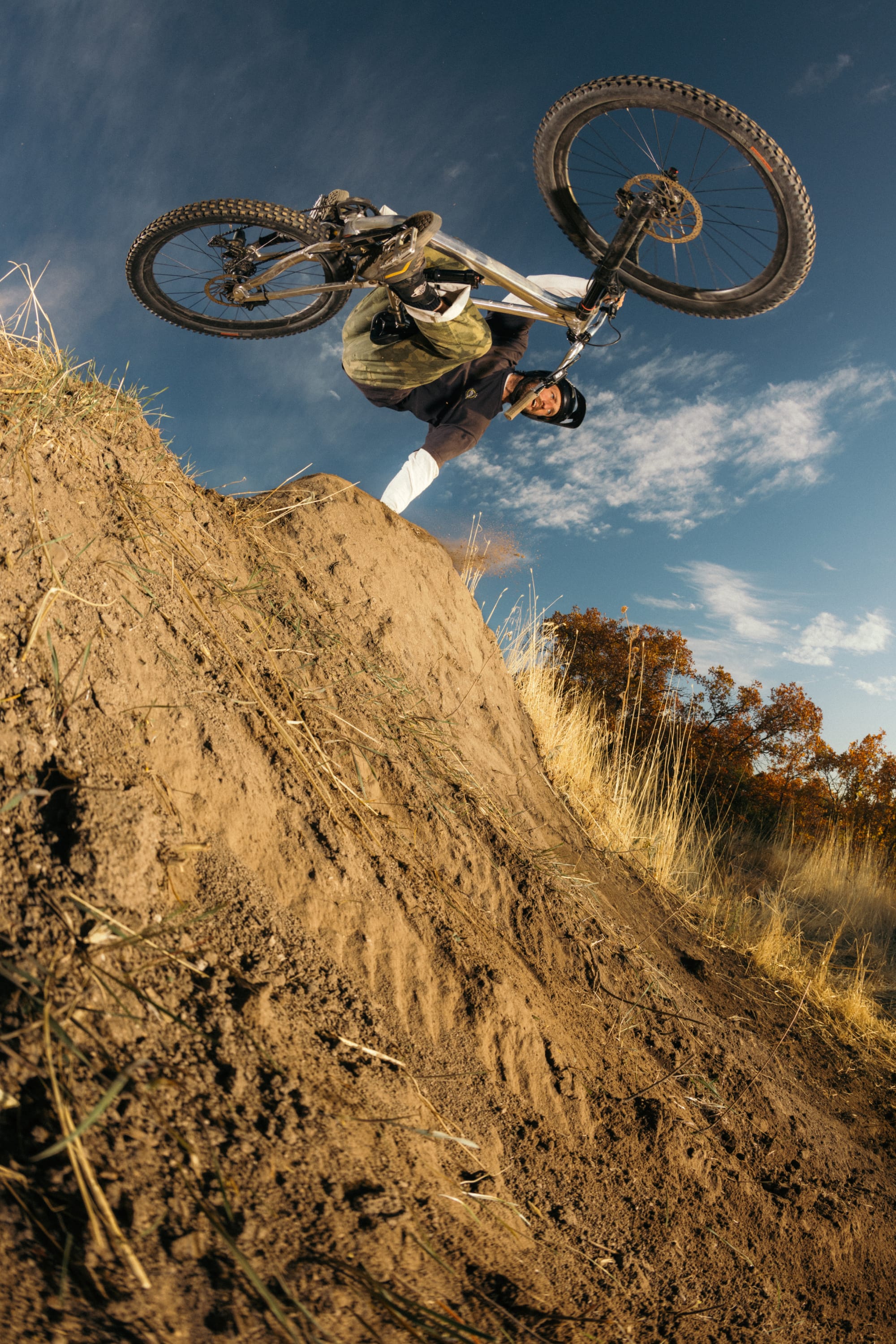 A mountain biker in a helmet and 686 Cruiser Pant - Wide Fit performs an aerial trick over a dirt ramp, tilting the bike sideways with blue sky and autumn foliage behind.