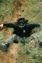 A person in a helmet, goggles, and 686 Cruiser Pant - Wide Fit by 686 sits partially inside a hole on a grassy, muddy hillside, arms outstretched and legs splayed.