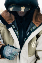 A person zips up their 686 Mens 2001 Transfer Jacket, paired with a camouflage cap, black hoodie, and camo-patterned gloves. The hat and hood mostly obscure their face.