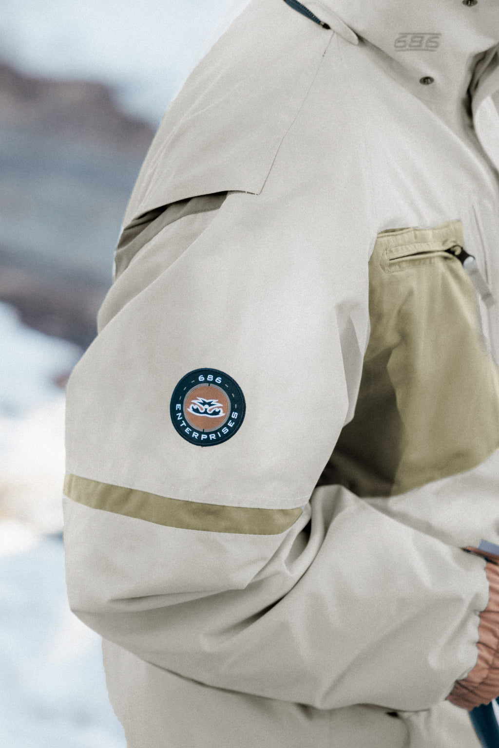 A person wearing the 686 Mens 2001 Transfer Jacket by 686, featuring a tan stripe and a circular GGG ENTERPRISES patch on the sleeve, stands outdoors in a snowy setting.