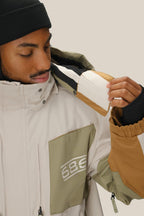 A man in a beige, green, and brown 686 Mens 2001 Transfer Jacket with a black beanie lifts the hood, revealing a white and brown patch inside. The waterproof polyester jacket features a zippered chest pocket with the white 686 logo.