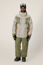 A person in a 686 Mens 2001 Transfer Jacket by 686 in beige and olive green, with matching pants, black gloves, dark boots, and ski goggles stands against a plain light background.