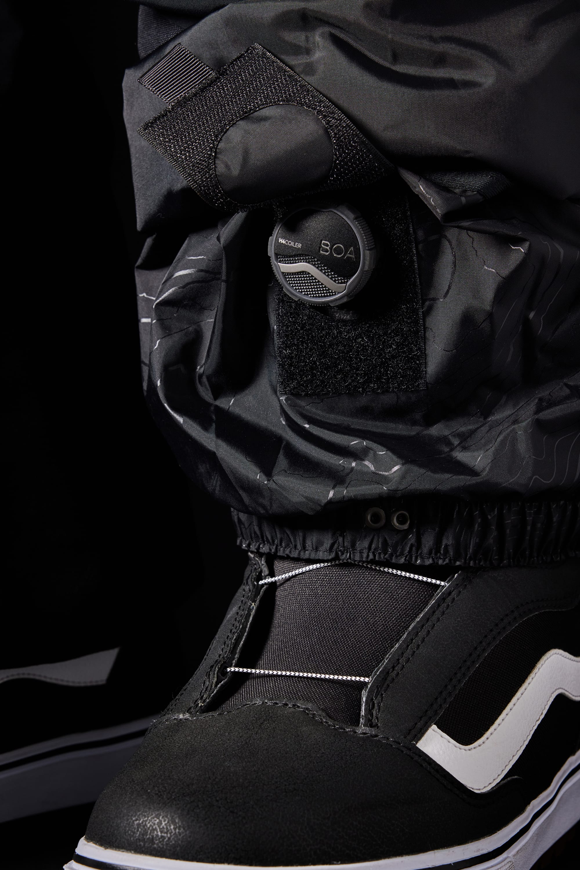 Close-up of a black sneaker with a white stripe, partly covered by the 686 Men's SMARTY® 3-in-1 Cargo Bib, with its shell pant leg secured by a round BOA tightening dial and Velcro strap.