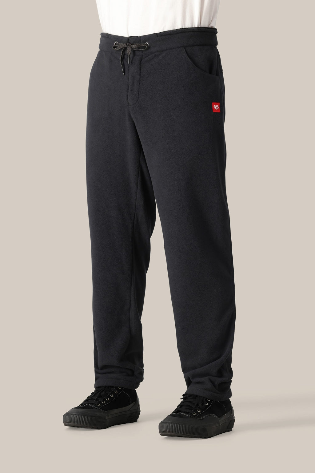 A person wearing the 686 Men's SMARTY® 3-in-1 Cargo Bib by 686, styled like a shell pant, with a black drawstring waist and red rectangular logo on the left pocket, paired with black sneakers and standing against a plain beige background.