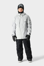 A person wearing a light gray hooded winter jacket, 686 Men's SMARTY® 3-in-1 Cargo Bib from 686, black gloves, snow boots, a black balaclava, and ski goggles stands against a plain light gray background.