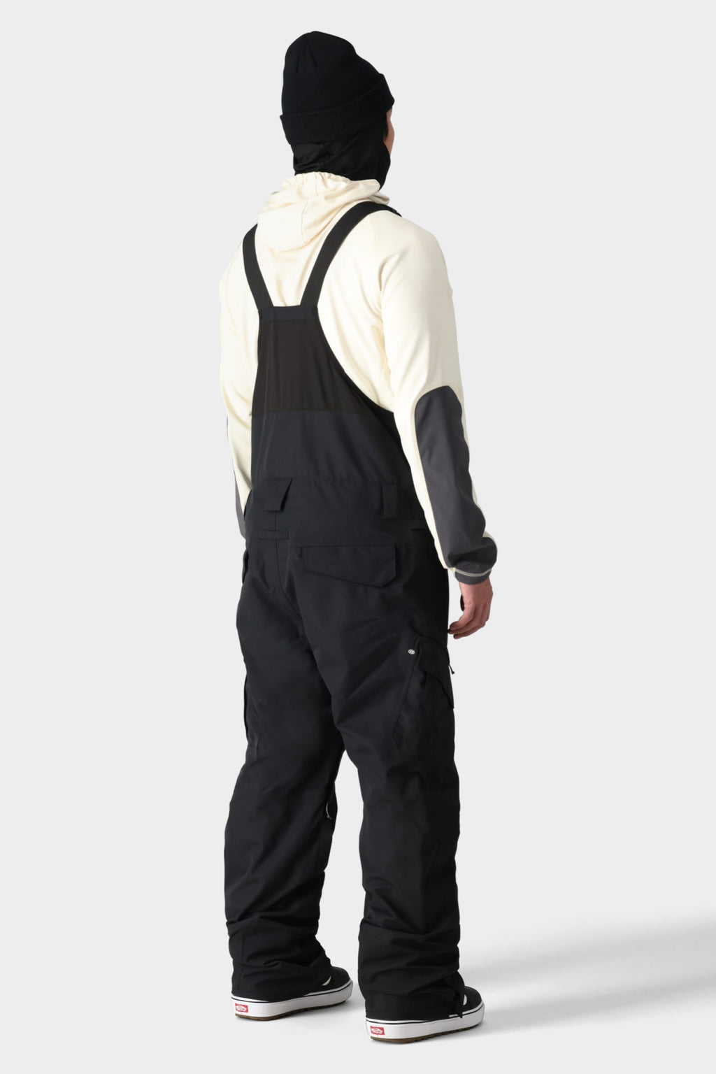 A person wearing a cream hoodie, black beanie, and 686 Men's SMARTY® 3-in-1 Cargo Bib stands with their back to the camera against a plain light background, showcasing the 686 brand snow overalls from behind.