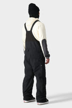 A person wearing a cream hoodie, black beanie, and 686 Men's SMARTY® 3-in-1 Cargo Bib stands with their back to the camera against a plain light background, showcasing the 686 brand snow overalls from behind.