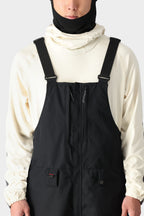 A person wears the 686 Men's SMARTY® 3-in-1 Cargo Bib overalls with adjustable straps, paired with a cream hoodie and black balaclava, standing against a light gray background.