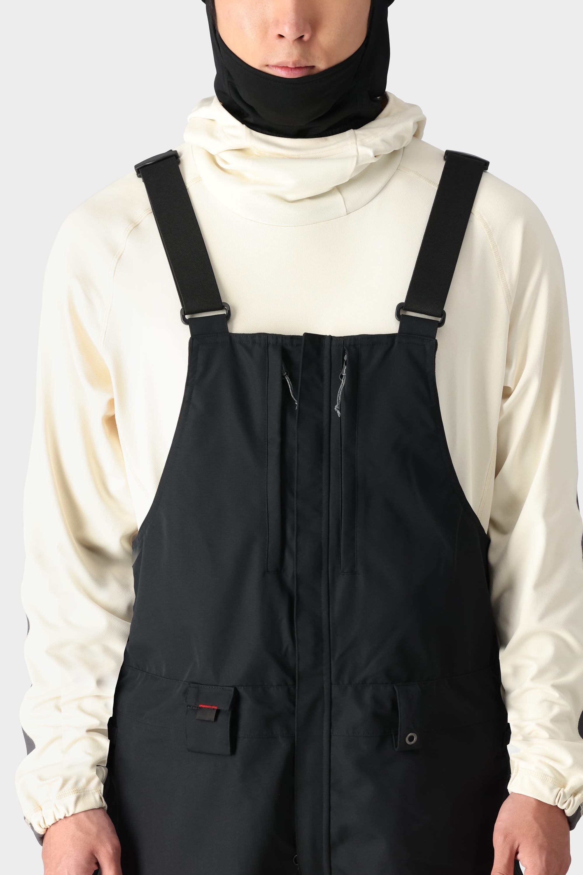A person wears the 686 Men's SMARTY® 3-in-1 Cargo Bib overalls with adjustable straps, paired with a cream hoodie and black balaclava, standing against a light gray background.