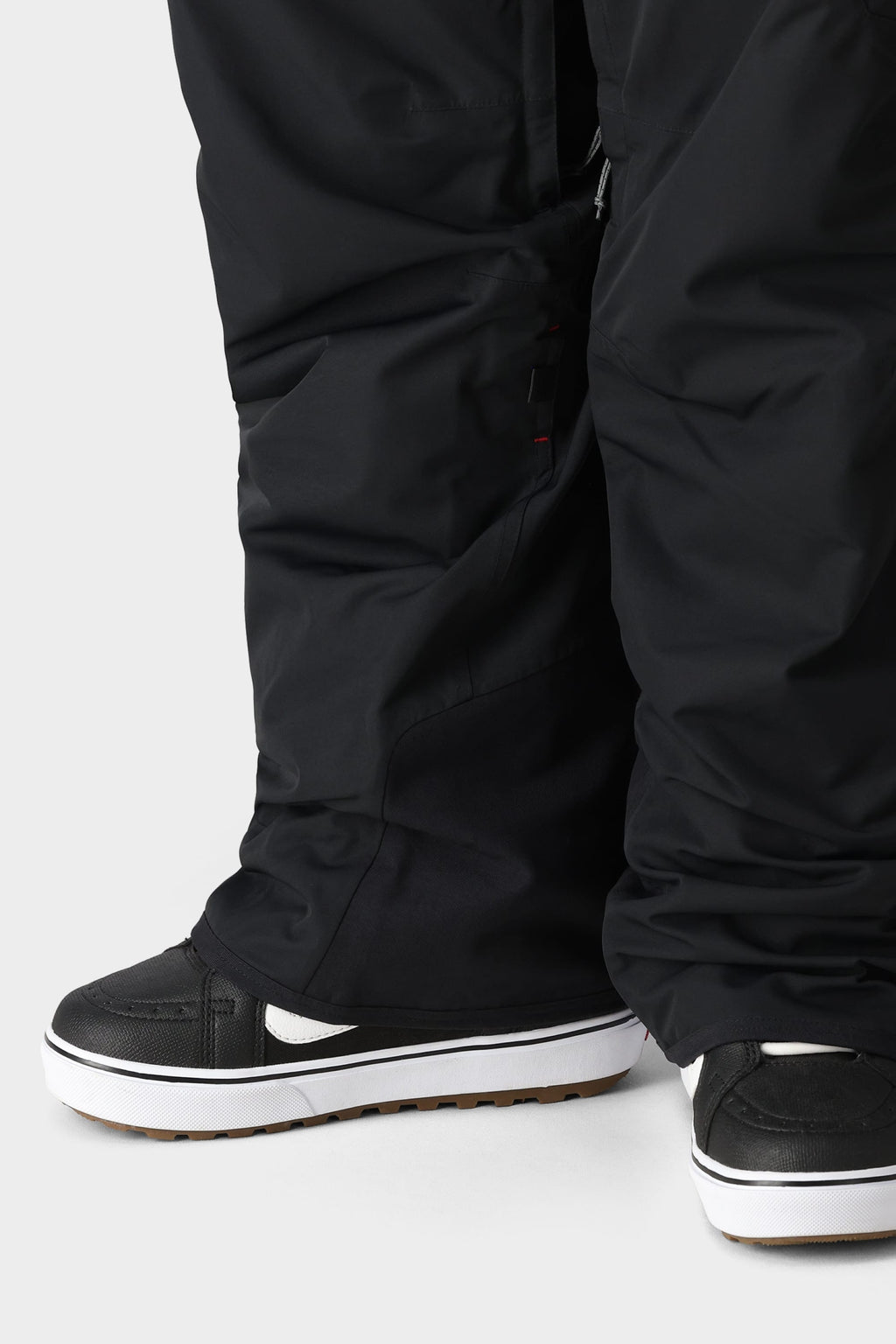 Close-up of a person wearing the 686 Men's SMARTY® 3-in-1 Cargo Bib, loose black shell pants from 686, partially covering black skate-style sneakers with white soles and brown tread, standing on a plain light background.