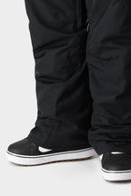 Close-up of a person wearing the 686 Men's SMARTY® 3-in-1 Cargo Bib, loose black shell pants from 686, partially covering black skate-style sneakers with white soles and brown tread, standing on a plain light background.