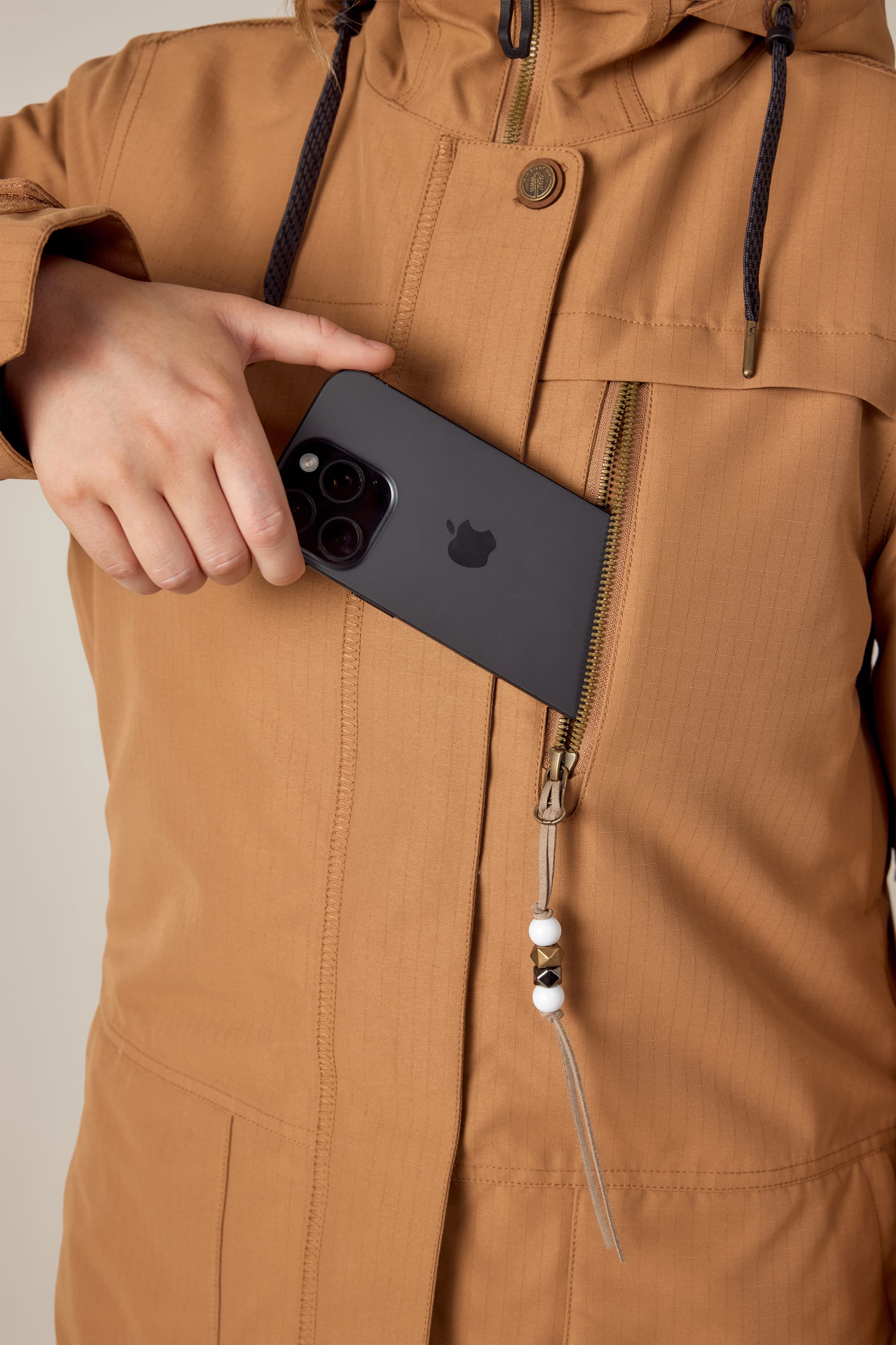 A woman wearing the 686 Womens Spirit Insulated Jacket in brown slips a black smartphone into the front zippered pocket.