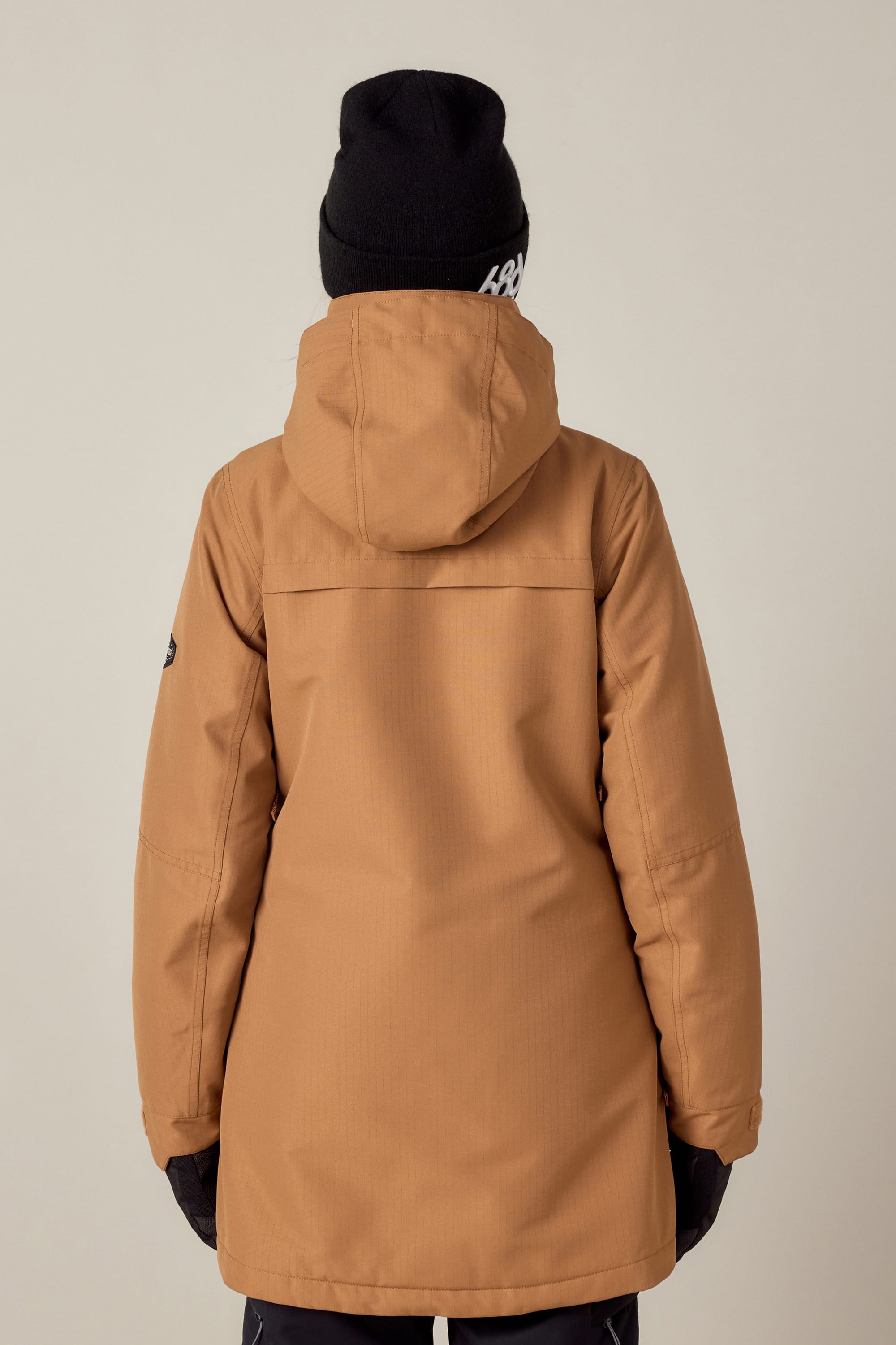 A person wearing the 686 Womens Spirit Insulated Jacket, a black beanie, gloves, and dark pants stands with their back to the camera against a plain light background.