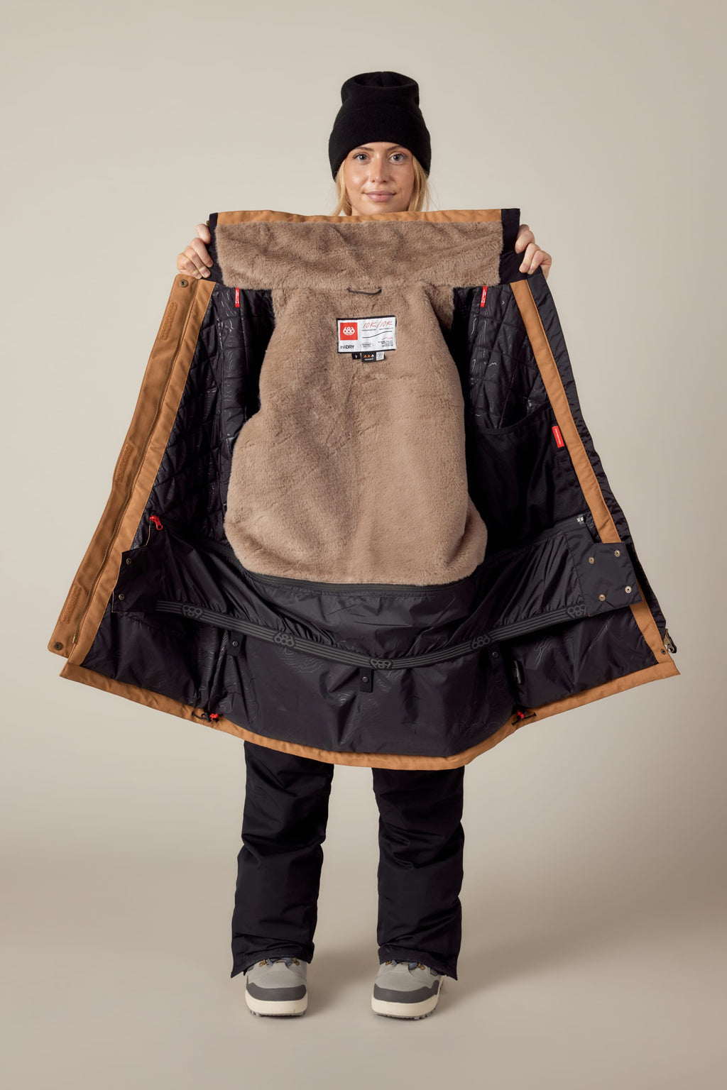 A smiling person in winter clothing and a black beanie holds open the 686 Womens Spirit Insulated Jacket, displaying its soft tan faux-fur lined interior and the 686 brand label.