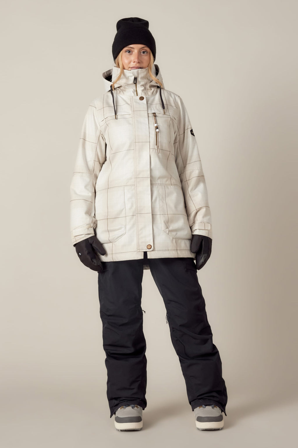 A person faces forward against a plain beige background, wearing the 686 Womens Spirit Insulated Jacket, black snow pants, black gloves, a black beanie, and winter boots.