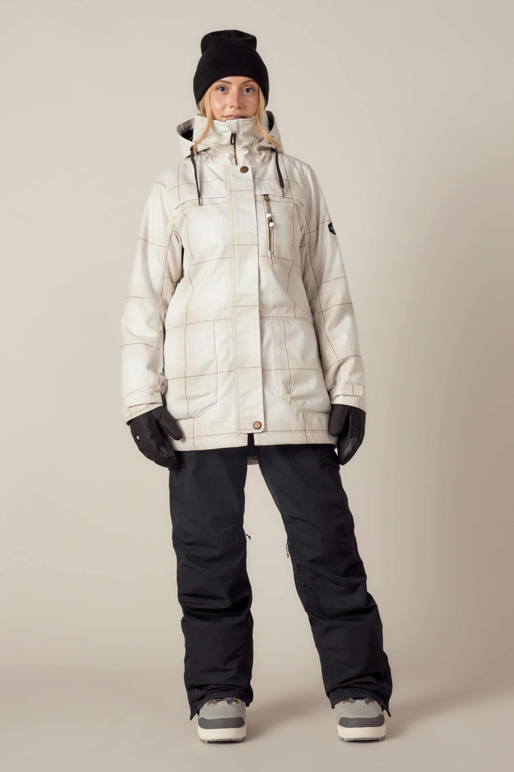 A person faces forward against a plain beige background, wearing the 686 Womens Spirit Insulated Jacket, black snow pants, black gloves, a black beanie, and winter boots.