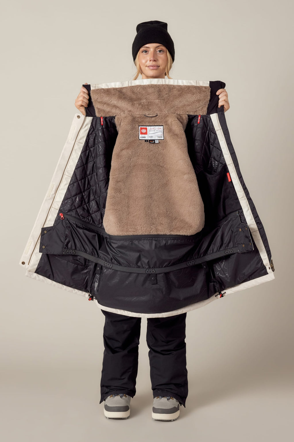 A person wearing snow gear holds open the 686 Womens Spirit Insulated Jacket, showing its warm brown and black faux-fur lined interior with tags and quilted sections.