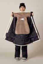 A person wearing snow gear holds open the 686 Womens Spirit Insulated Jacket, showing its warm brown and black faux-fur lined interior with tags and quilted sections.