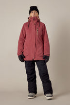 A person stands facing forward against a plain beige background, wearing the 686 Womens Spirit Insulated Jacket in red by 686, paired with a black beanie, black gloves, black snow pants, and gray winter boots.