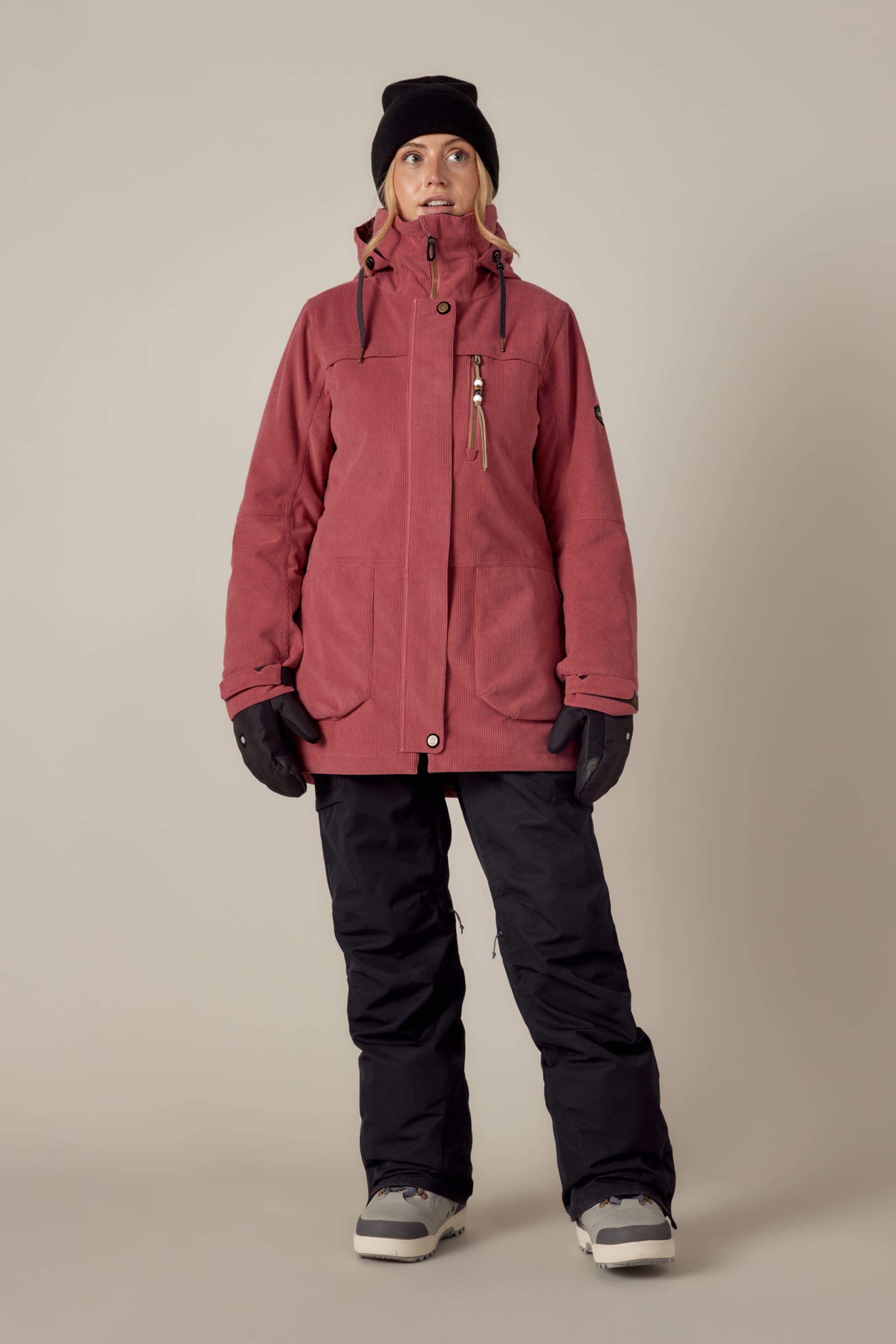 A person stands facing forward against a plain beige background, wearing the 686 Womens Spirit Insulated Jacket in red by 686, paired with a black beanie, black gloves, black snow pants, and gray winter boots.
