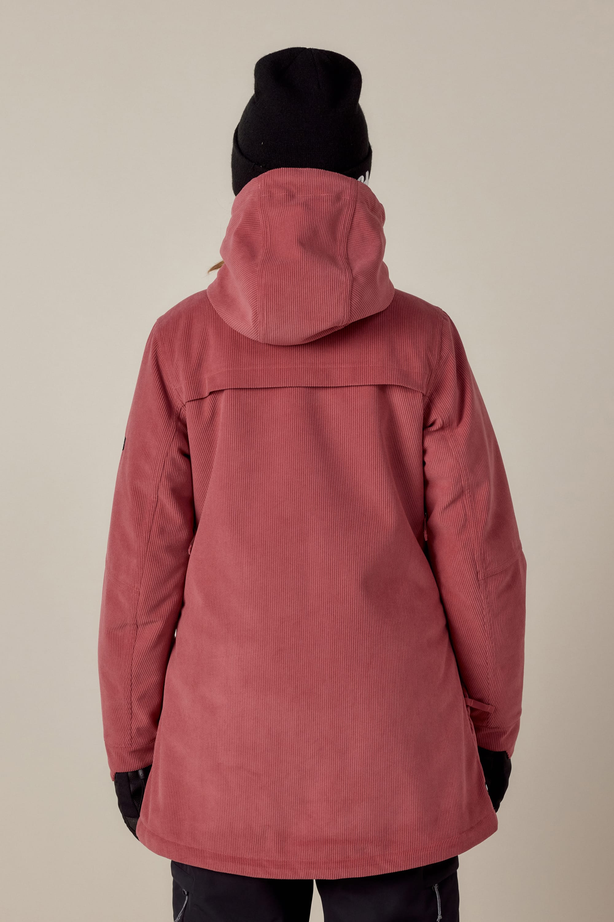 A person is shown from the back against a plain light background, wearing the 686 Womens Spirit Insulated Jacket by 686 in dusty rose with faux-fur lined hood, paired with a black knit beanie, black gloves, and dark pants.