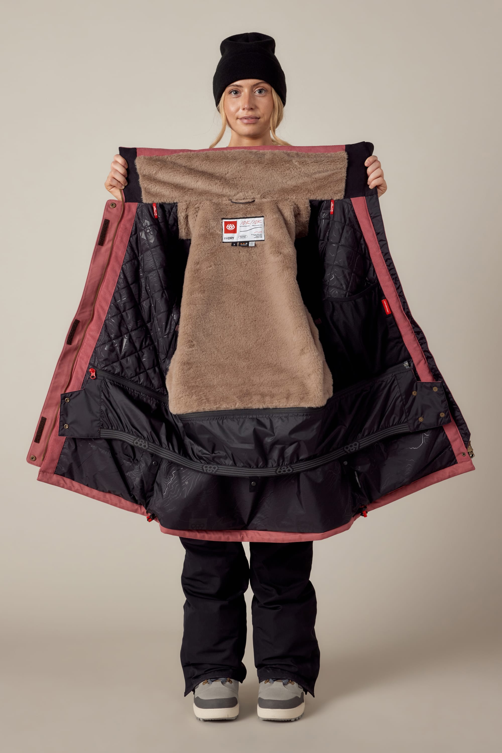A person in winter clothing opens a maroon and black 686 Womens Spirit Insulated Jacket, showing the soft tan and quilted black lining. They also wear black pants, gray shoes, and a black beanie, standing against a plain background.
