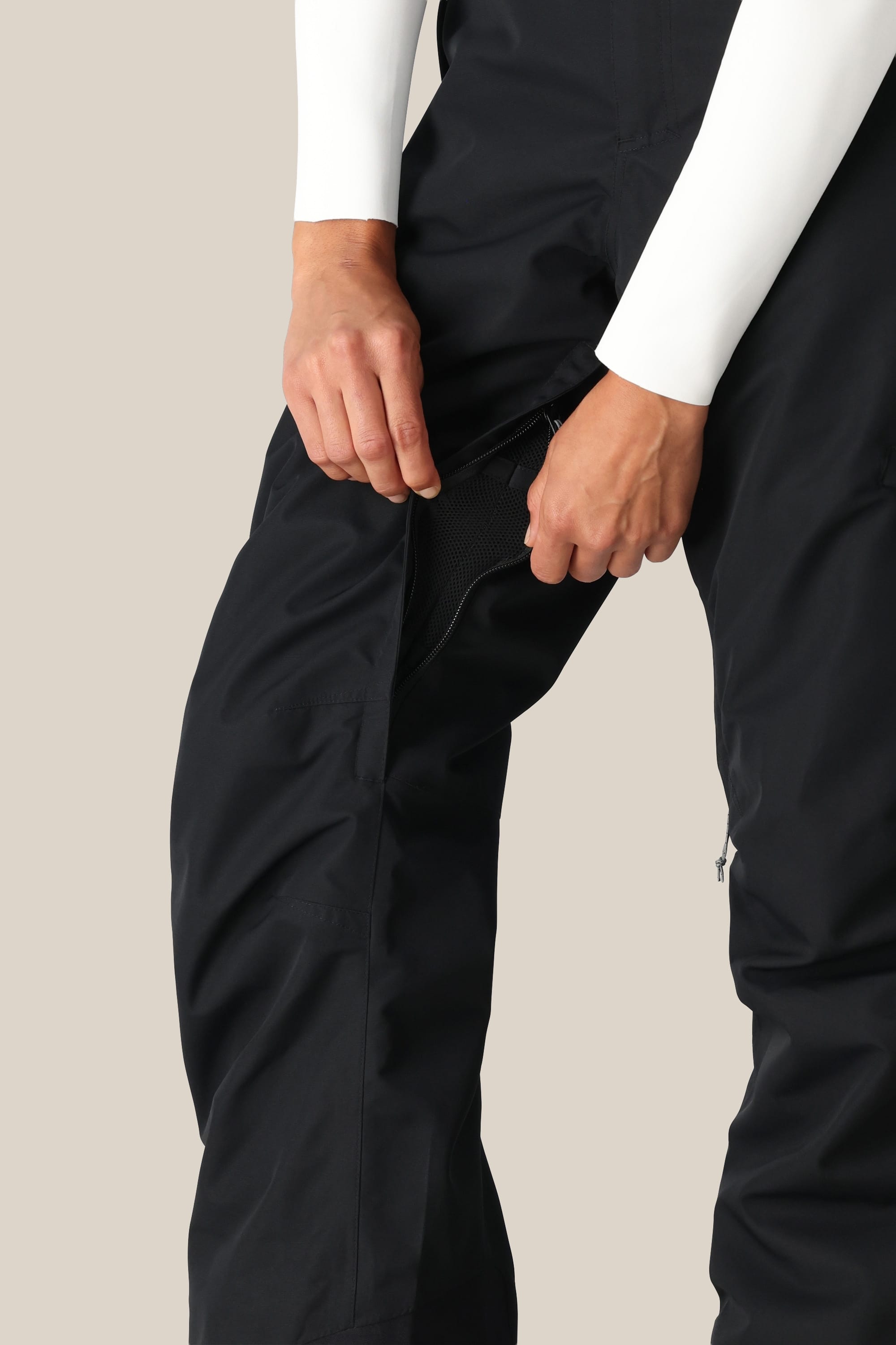 A person wearing the 686 Women's SMARTY® 3-in-1 Cargo Pant unzips a ventilation zipper on the side. The image highlights their lower torso and hands against a neutral beige background.