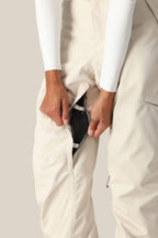 A person in 686 Women's SMARTY® 3-in-1 Cargo Pant and a white long-sleeve top unzips a mesh-lined side vent. These cargo pants feature a removable sweatpant liner for versatile wear. The background is plain and neutral.