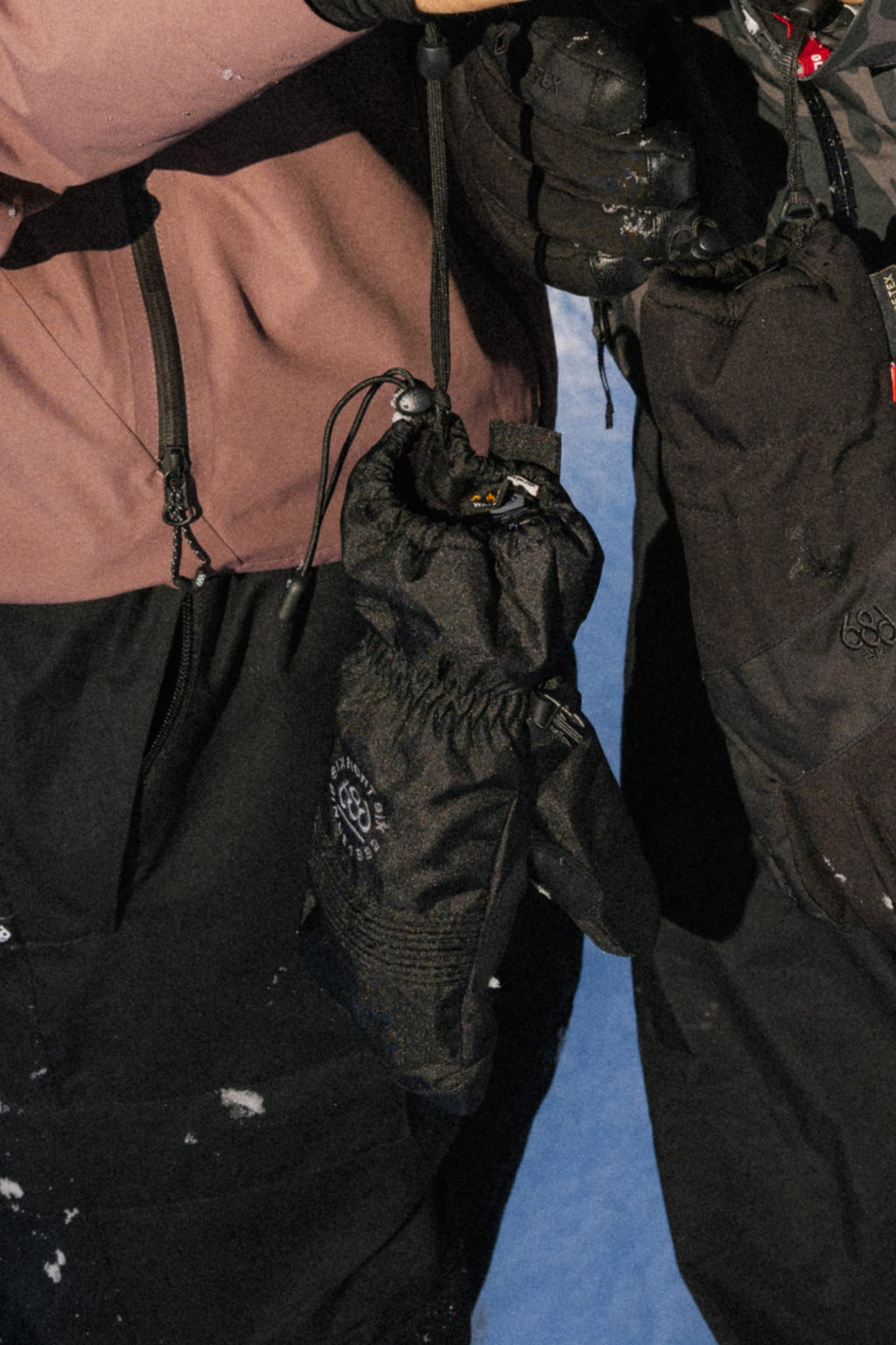 Two people wearing 686 Lander™ Mitts by 686, along with winter jackets, stand close together holding the drawstrings of their black gloves. Snow covers their clothing and the ground beneath them.