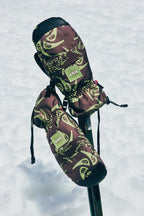 Two patterned 686 Lander™ Mitts with light insulation and waterproofing, featuring the 686 logo, are placed atop crossed ski poles against a snowy backdrop.
