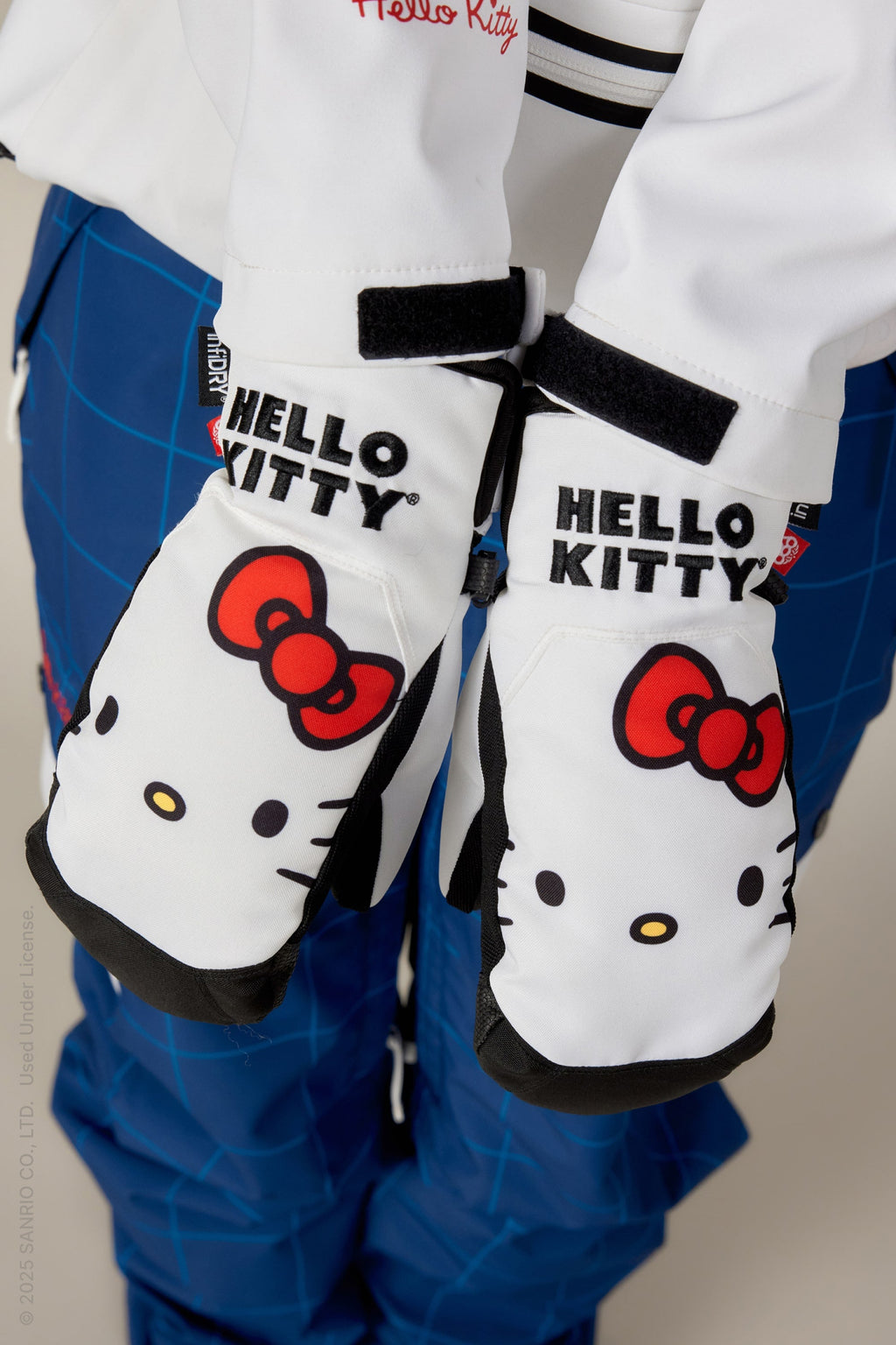 A person wearing 686 Womens Revel Mitts by 686—white waterproof mittens with Hello Kitty, red bows, and HELLO KITTY printed on each mitten—paired with blue plaid pants.