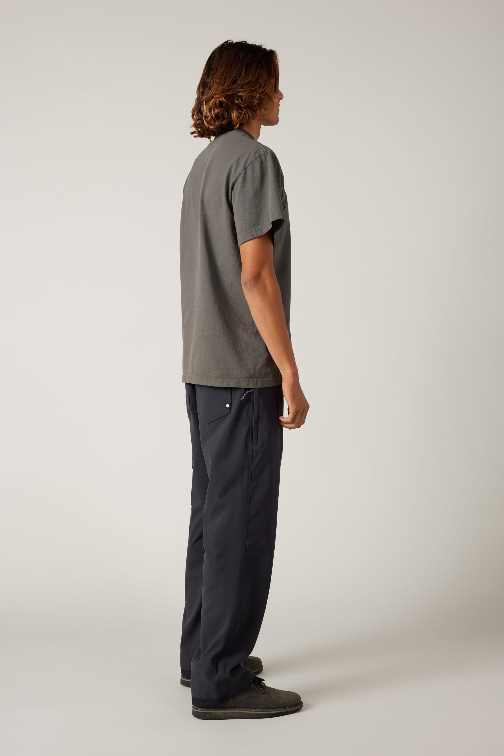 A person with shoulder-length hair stands sideways, wearing a loose gray t-shirt and 686 Men's Unwork® Pant - Relaxed Loose Fit, paired with dark shoes, against a plain light background.